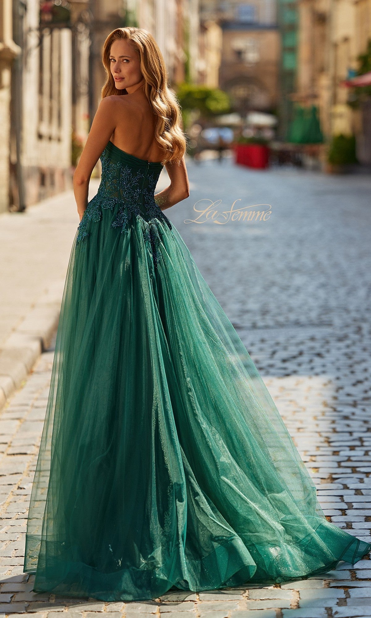 A woman stands on a cobblestone street, looking over her shoulder, wearing the La Femme 33528 long prom dress featuring a strapless sheer corset bodice and a floor-length emerald green skirt.